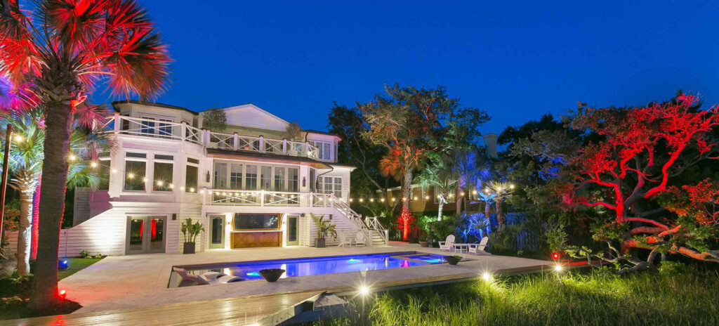 A brightly lit modern house with large windows and a spacious deck overlooks a swimming pool, enhanced by an outdoor lighting franchise, surrounded by trees and colorful lights at night.