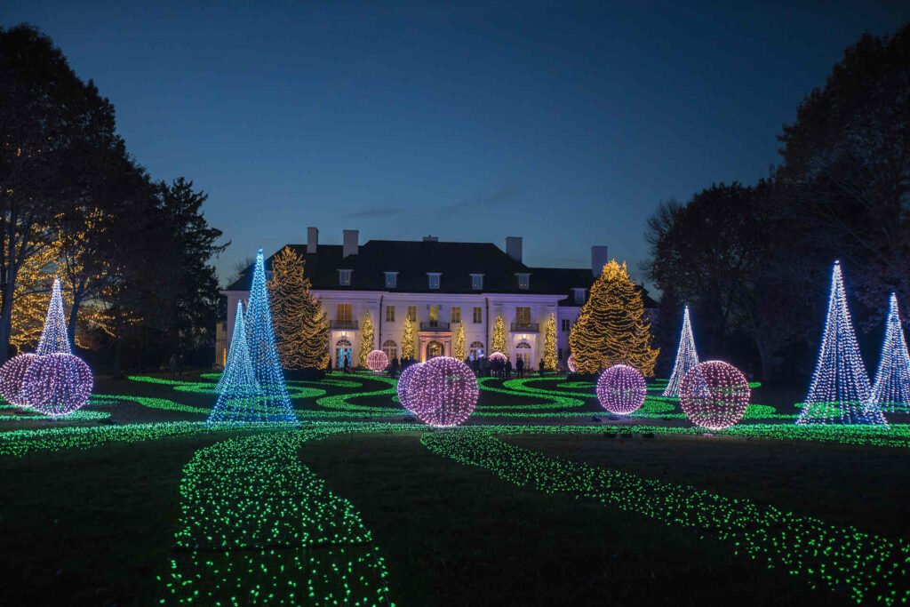 A large mansion is illuminated at night with decorative lights, including glowing cones, spheres, and green paths, set in a landscaped garden.
